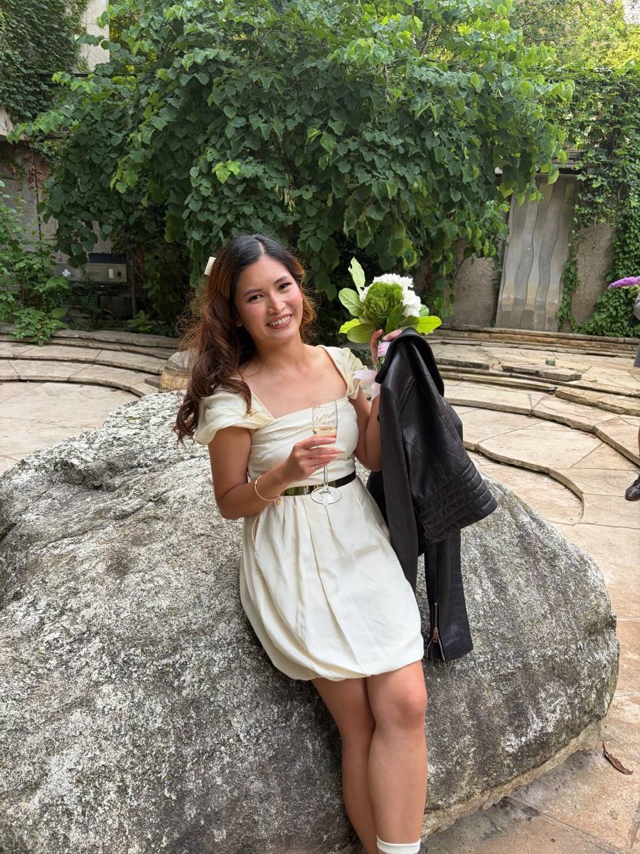 Joelle in the vintage dress, holding a bouquet and a glass, seated on a rock in a stone courtyard